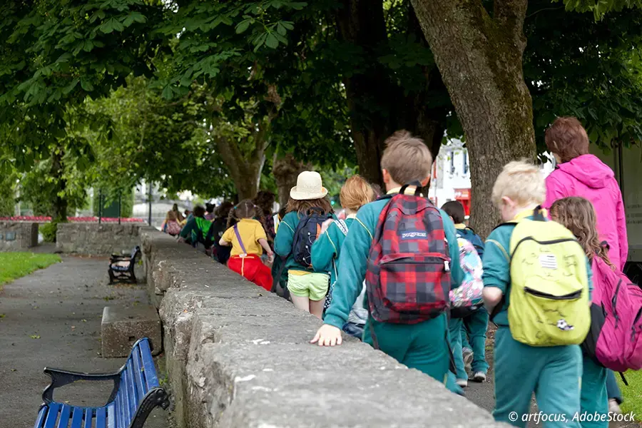 Eine Gruppe von Schulkindern mit Rucksäcken spaziert durch eine grüne städtische Parklandschaft.